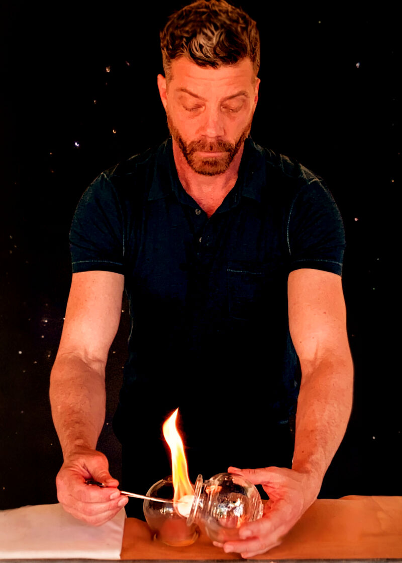 Dr. Bill Bastian igniting a cotton wick inside a glass cupping jar, dark backdrop
